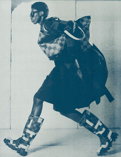 Fashion shot of an adult Black male model wearing sunglasses, coat, skirt, purse, and rain boots with a geometric pattern in a studio.