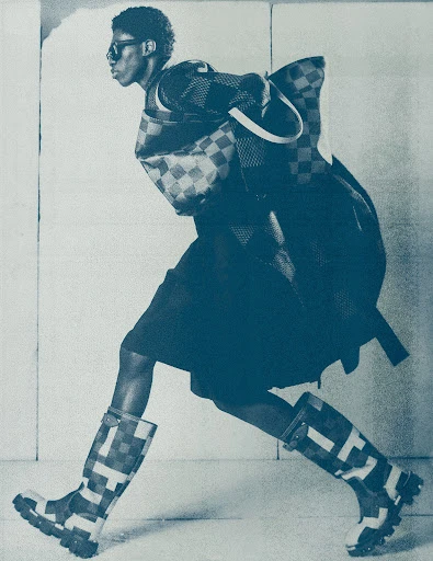 Fashion shot of an adult Black male model wearing sunglasses, coat, skirt, purse, and rain boots with a geometric pattern in a studio.