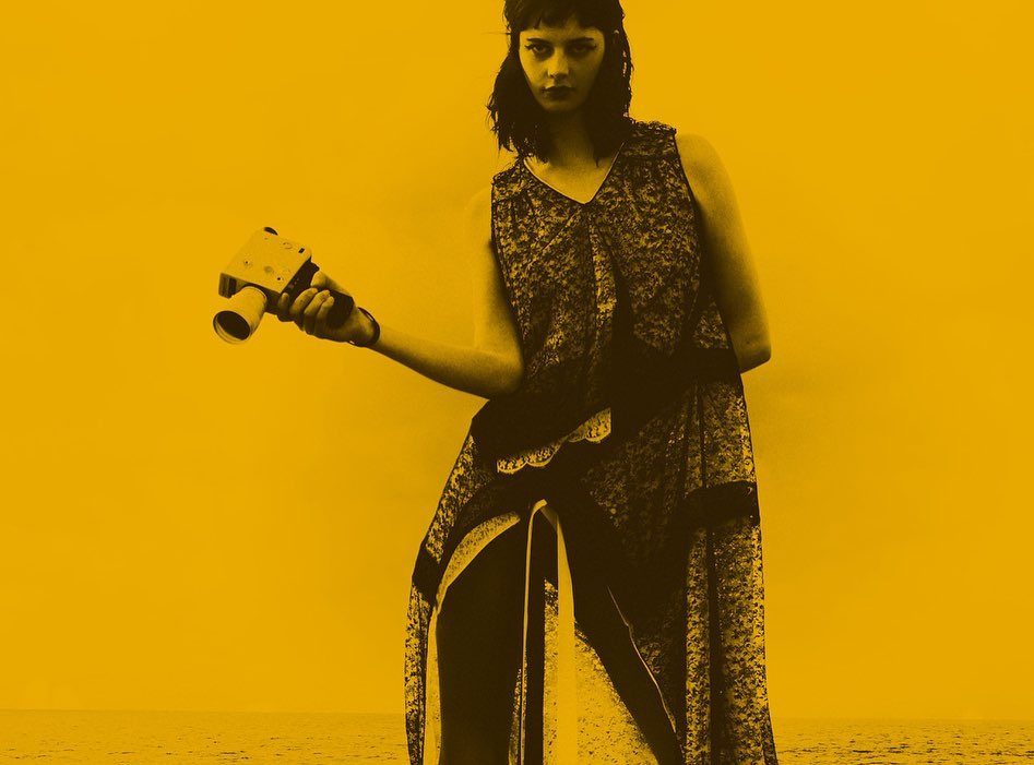Woman poses outdoors in long dress with retro camera, against yellow background and ocean horizon in fashion-style photography.