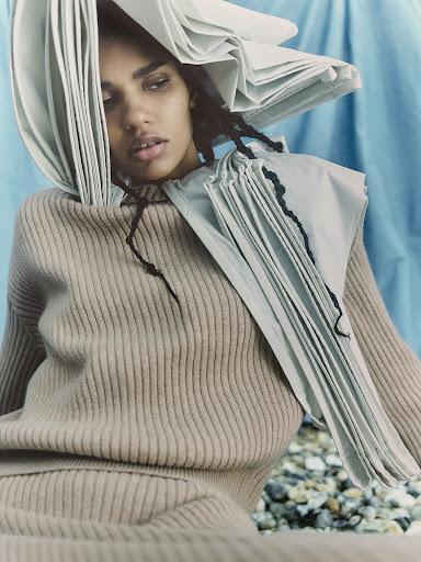 Fashion portrait of a person with medium brown skin, wearing a sweater and a sculptural paper headdress in a studio setting.