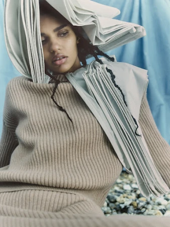 Fashion portrait of a person with medium brown skin, wearing a sweater and a sculptural paper headdress in a studio setting.