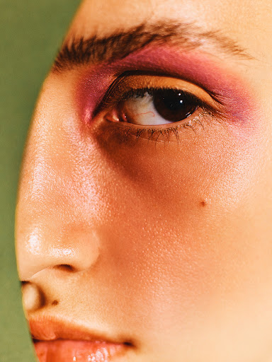 Close-up of a woman's face featuring pink eyeshadow. The shot showcases her eye, eyebrow, cheek, nose, and lips against a green background.