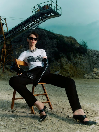 A fashion shot presents a woman in sunglasses, T-shirt, black gloves, and pants seated on a chair in a quarry setting with machinery.