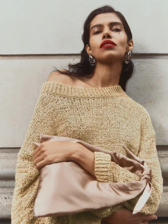 Brunette woman poses wearing a beige knit sweater and holding a tan satin clutch bag with a beige wall in the background.