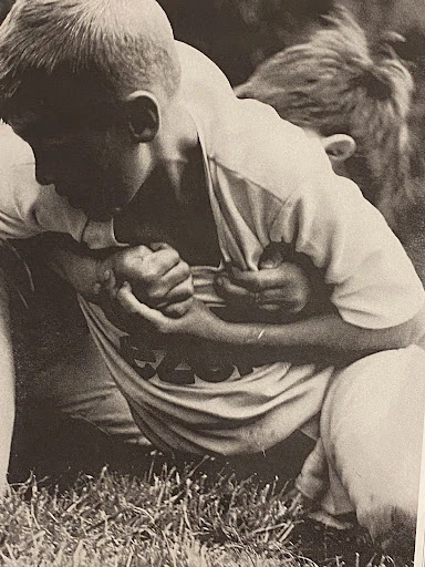 Black and white image of two boys wrestling on grass, one in a light-colored t-shirt with a graphic, outdoors during daytime.