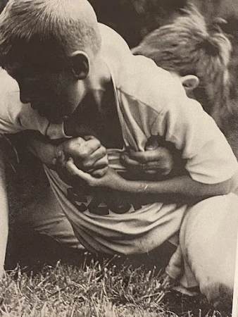 Black and white image of two boys wrestling on grass, one in a light-colored t-shirt with a graphic, outdoors during daytime.