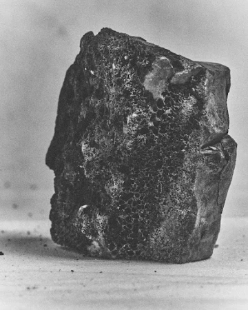 Monochrome close-up photograph of a rough textured dark mineral specimen resting on a flat surface