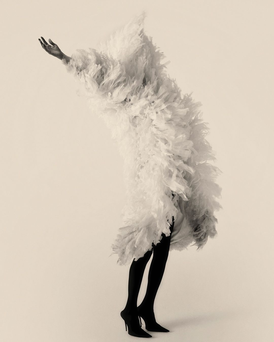 Monochrome fashion shot: person draped in textured white fabric, dark leggings, and stiletto heels against a neutral background.