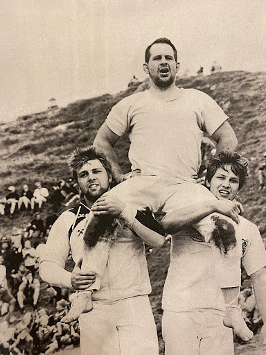 Three men in vintage photograph. One man stands on the shoulders of two other men against a hillside background with seated crowd.