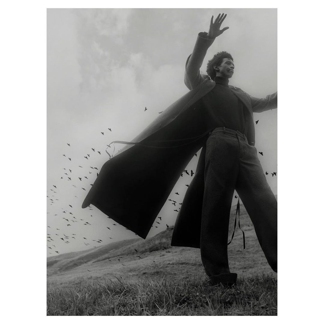 Monochrome portrait of a stylish African American man in a long coat, standing in a field with birds and sky in the background.