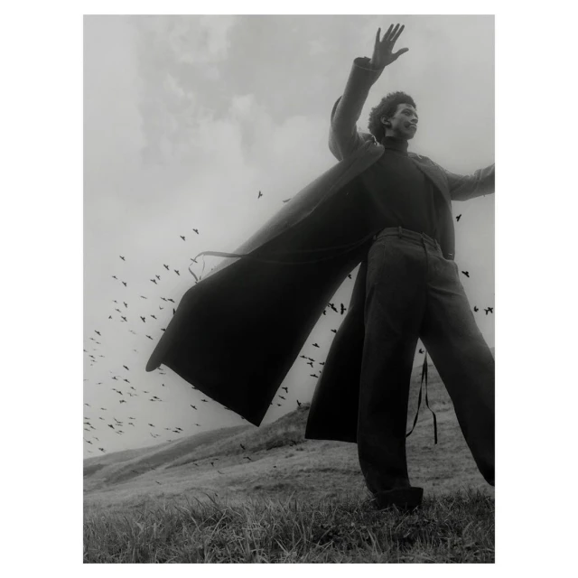 Monochrome portrait of a stylish African American man in a long coat, standing in a field with birds and sky in the background.