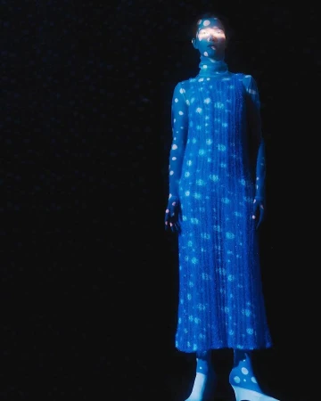 Model standing in darkness wearing a blue patterned dress and tights, illuminated by projected light spots