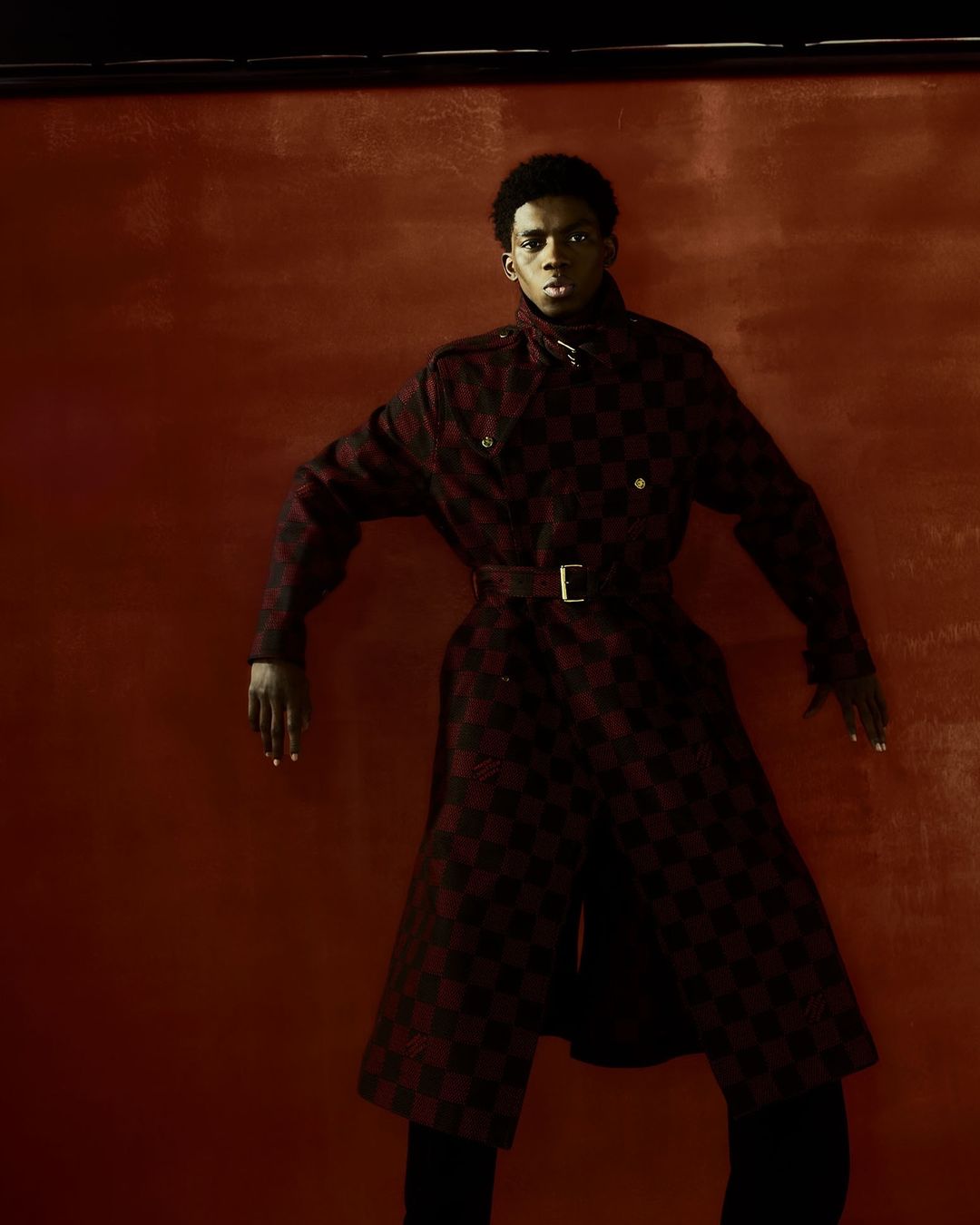 Fashionable Black male model in a patterned overcoat poses in front of a painted red-brown backdrop in a studio environment.