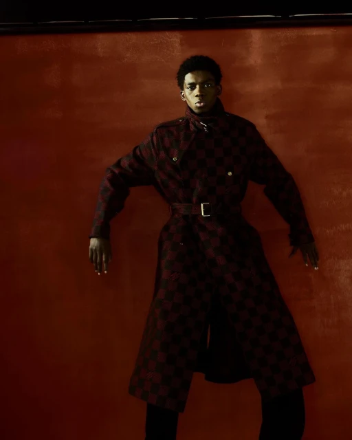 Fashionable Black male model in a patterned overcoat poses in front of a painted red-brown backdrop in a studio environment.