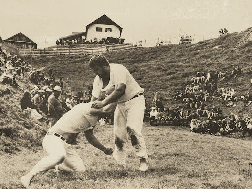 Two men wrestling in an outdoor arena, captured in a vintage photo, showcasing a crowd watching from a grassy hillside on a cloudy day.