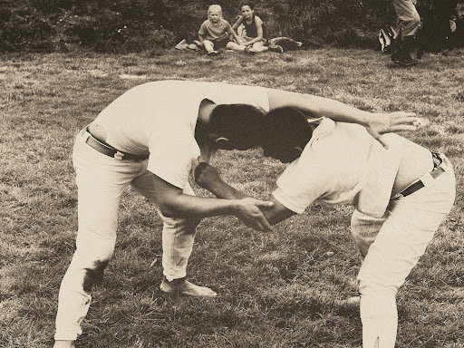 Two men wrestling barefoot in a grassy field, with two seated children visible in the background on a sunny day.