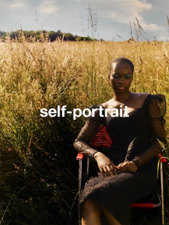 Black woman seated in a red folding chair wearing a dark sheer dress amidst tall meadow grass under an open sky