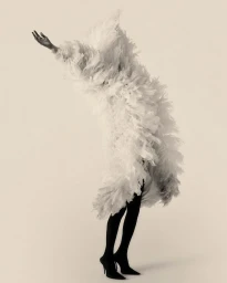 Model fully enveloped in a voluminous white feather coat raising one hand against a pale studio background