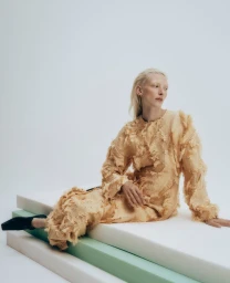 Model with blonde hair wearing a textured fringed beige outfit seated on stacked foam blocks against a clean background