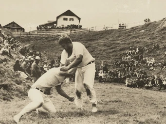 Two men wrestling in a grassy arena surrounded by spectators with a building on a hill in the background