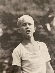 Portrait of a young blond boy wearing a light-colored t-shirt against a dark foliage background by Nicolas Kern