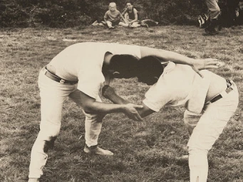 Two men wrestling on grass outdoors with children watching in the background, captured in a vintage monochrome style.