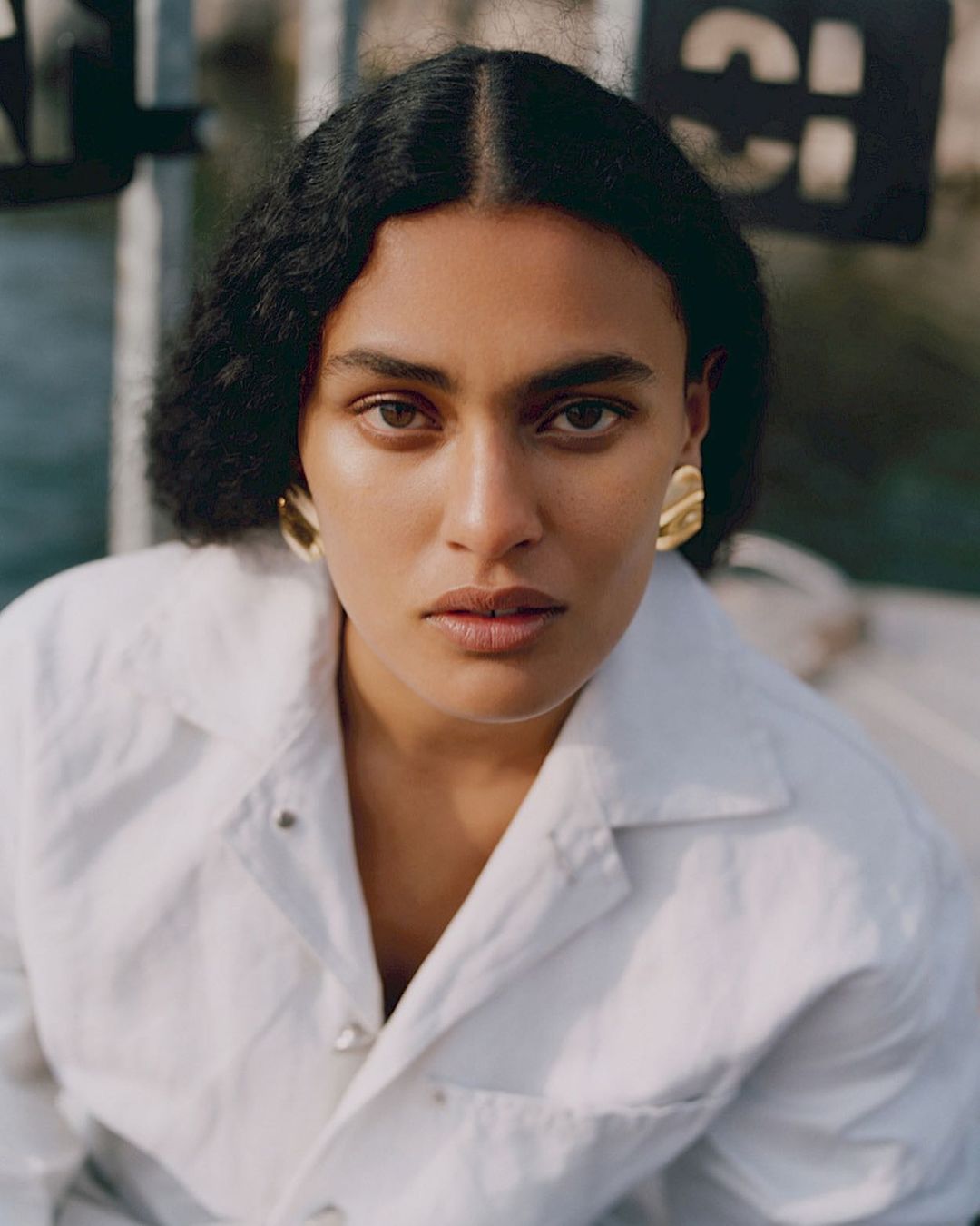 Close-up portrait of a brunette woman with large gold earrings wearing a white button-down shirt. The background is blurred and neutral.