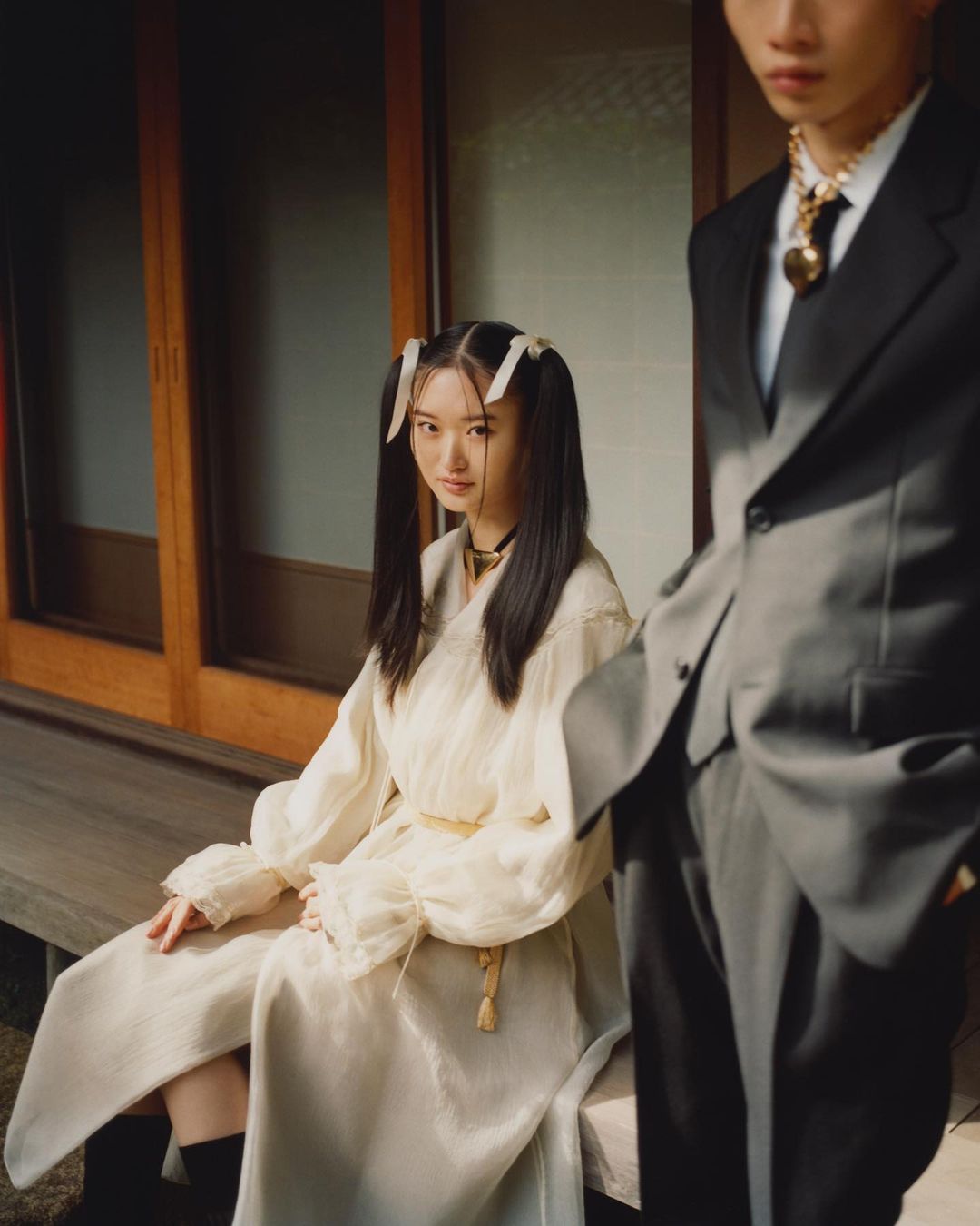 Two young adults in formal wear pose in a Japanese-style interior with neutral colors, creating a fashion editorial image.