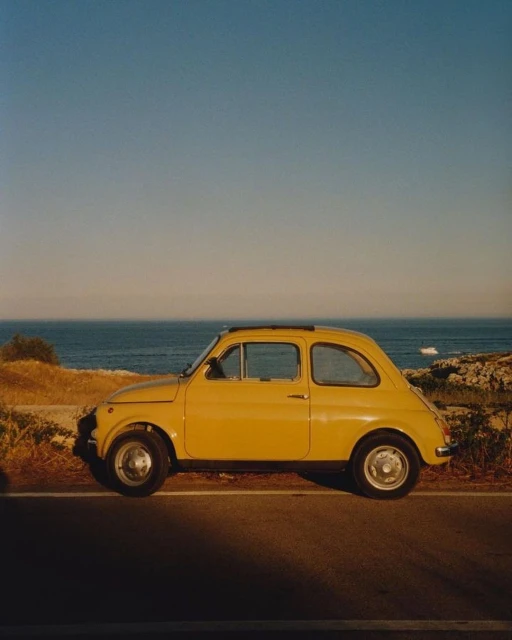 Yellow vintage car parked on a roadside near the ocean. The vehicle is bathed in warm light under a clear blue sky.
