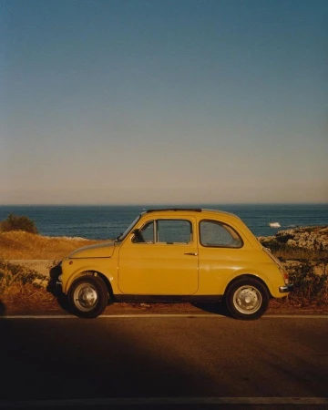 Yellow vintage car parked on a roadside near the ocean. The vehicle is bathed in warm light under a clear blue sky.