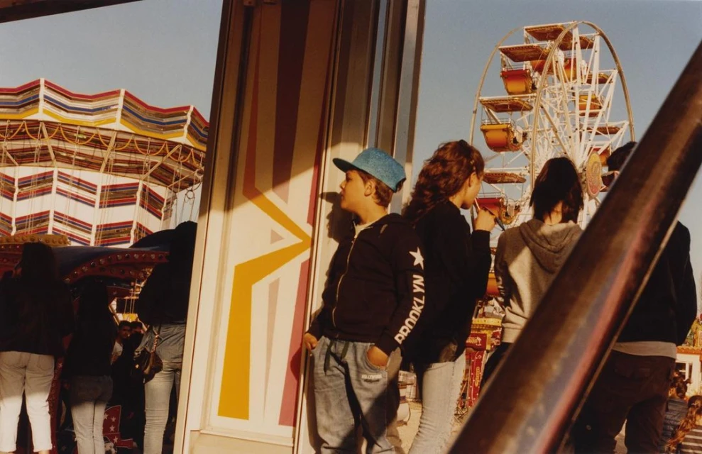 Young people at an outdoor amusement park with a Ferris wheel and ride, captured in natural light with a close-up shot.