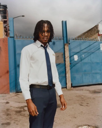 Portrait of a young adult male with dreadlocks wearing a white dress shirt and tie, standing in front of a blue metal gate outdoors.