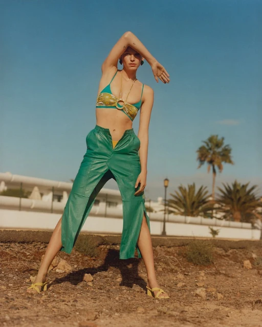 A woman poses in a bikini top and green pants outside against a clear blue sky with palm trees in the background, creating a sunny fashion shot.