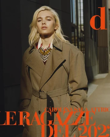 Portrait of a blonde woman wearing a belted beige trench coat and patterned sweater under natural sunlight outdoors.