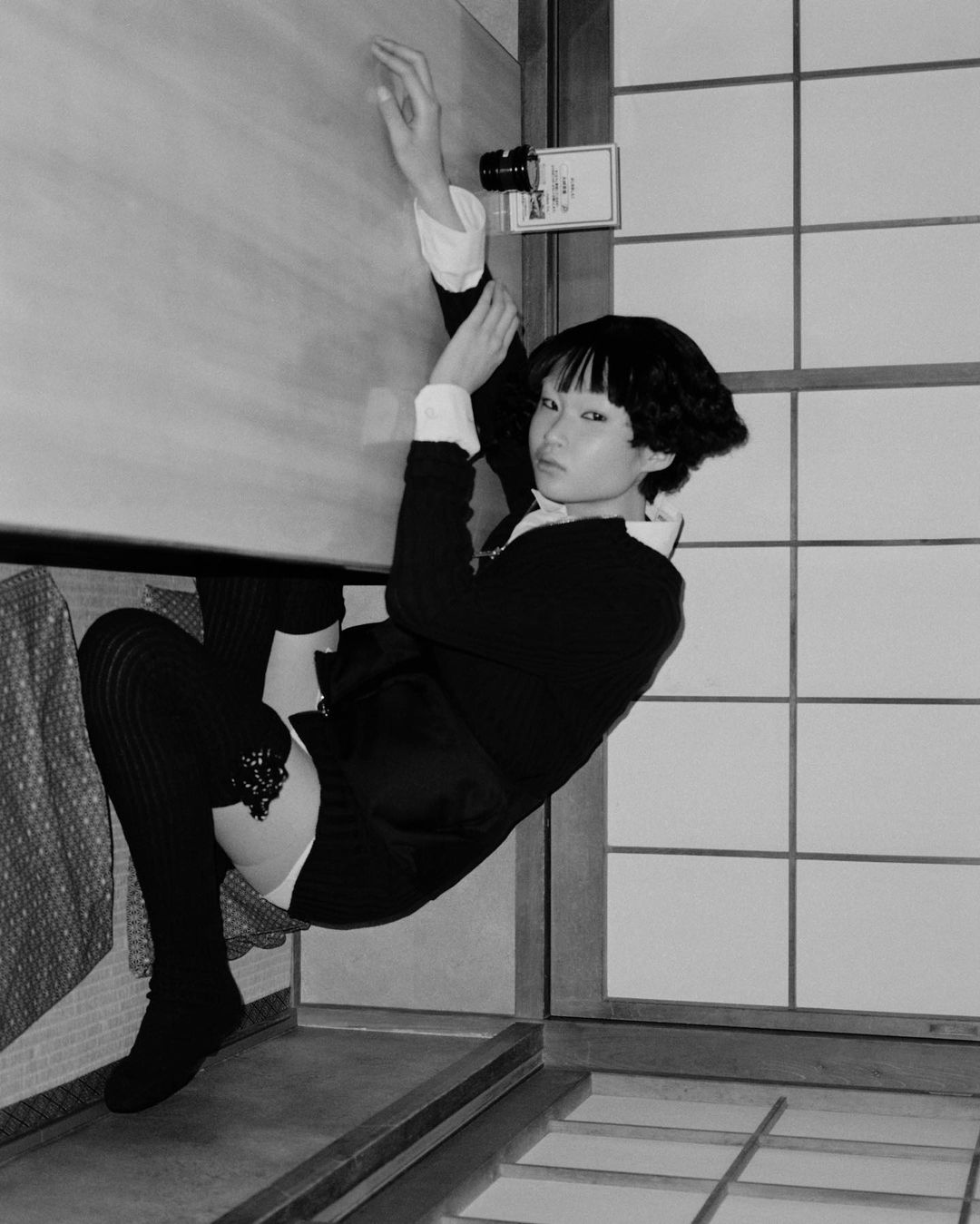 An Asian woman with a bob haircut poses indoors, wearing a black and white shirt, dark skirt, and knee-high socks in a corner studio.