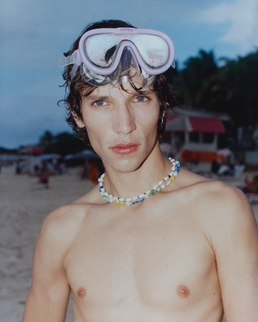 Young adult with swimming goggles on forehead and beaded necklace poses shirtless on beach with blurred figures and buildings in background.