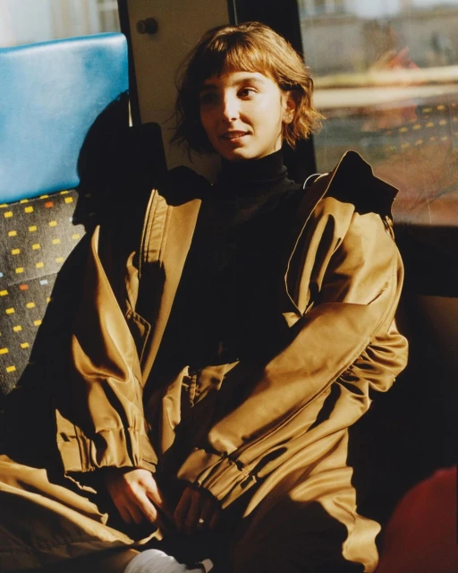 Young woman with short hair and brown jacket posing inside vehicle; warm, natural lighting.