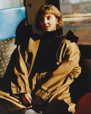 Young woman with short hair and brown jacket posing inside vehicle; warm, natural lighting.