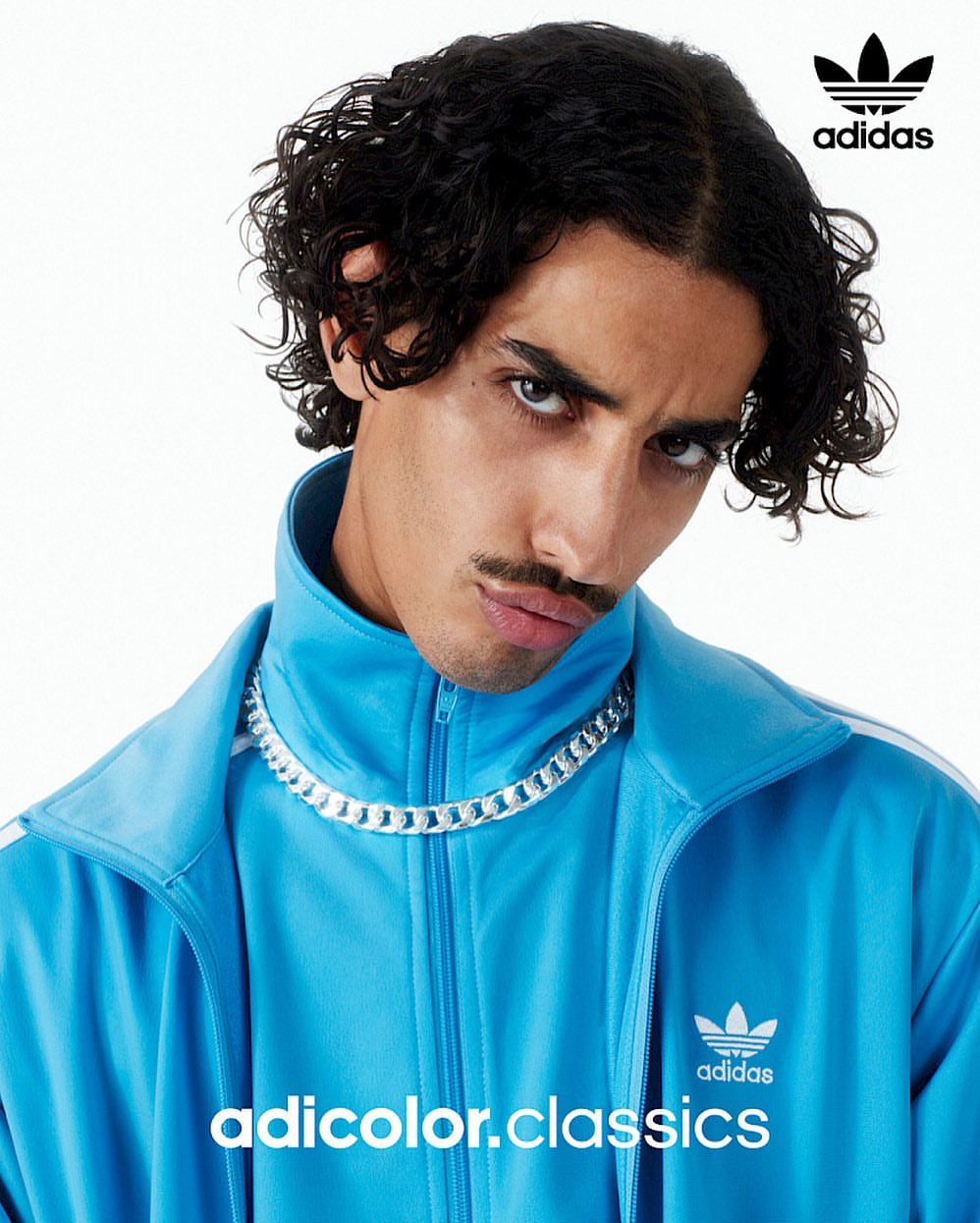 Close-up portrait of a young adult male model with dark curly hair, wearing a light blue Adidas tracksuit and silver chain necklace.