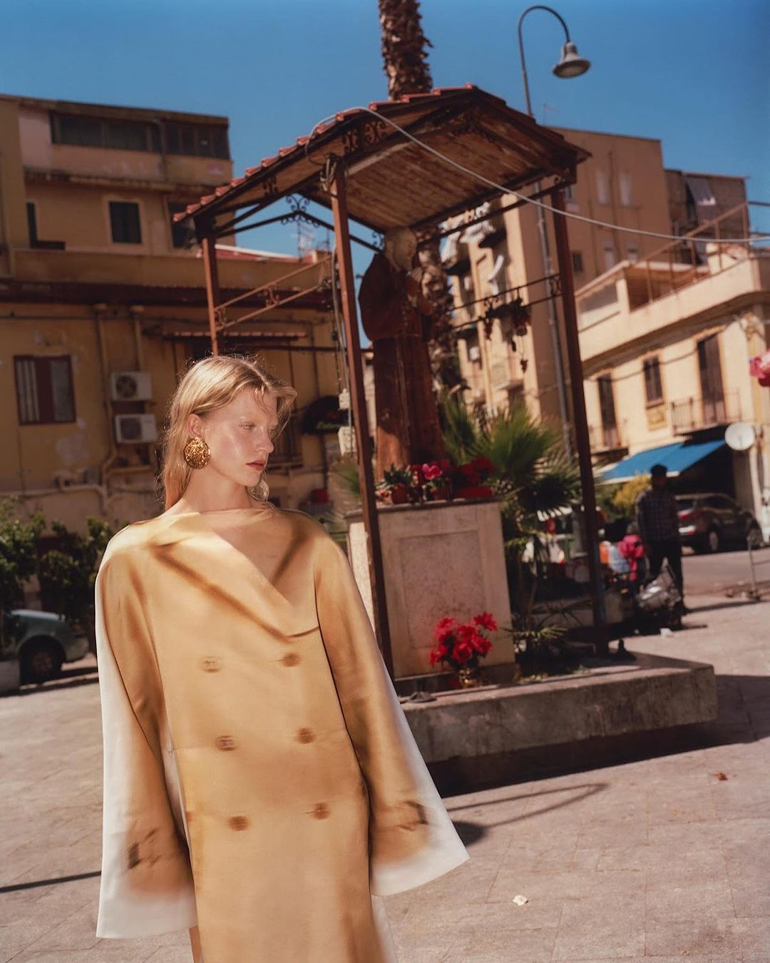 Fashion editorial shot of a blonde woman in a tan coat standing in a town square with buildings in the background.