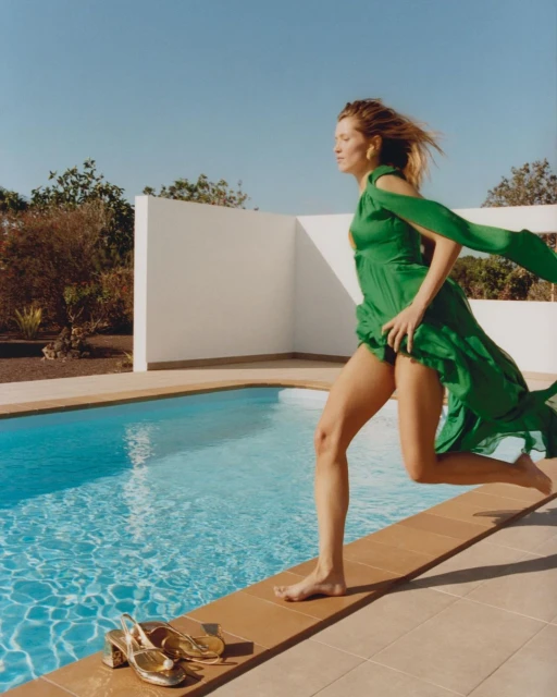 Woman in green dress barefoot near pool with golden sandals. Clear blue sky above a wall, plants, and tiled patio on a sunny day.