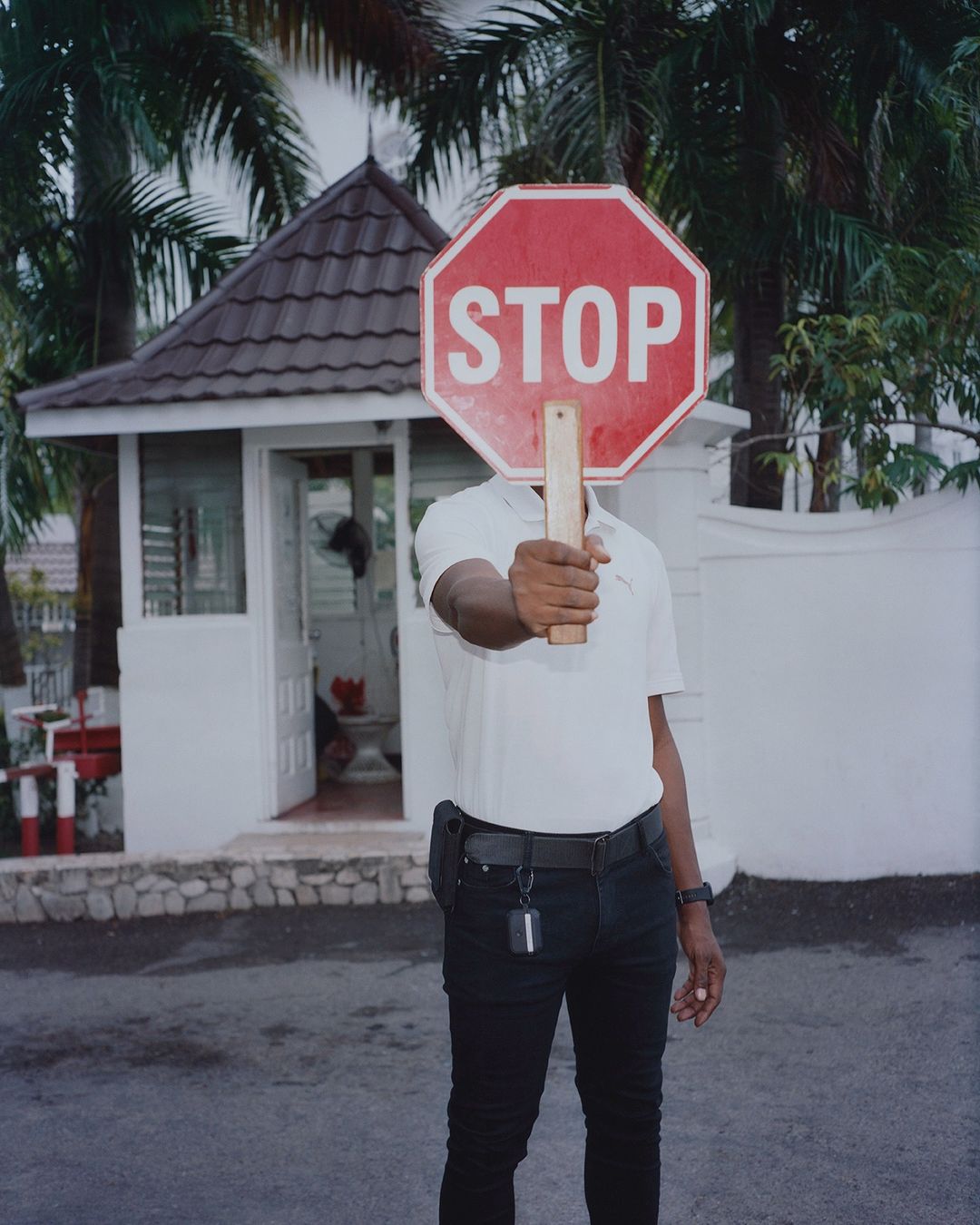 Person holds red stop sign with white letters in front of their face on an outdoor street near a white building and green trees.