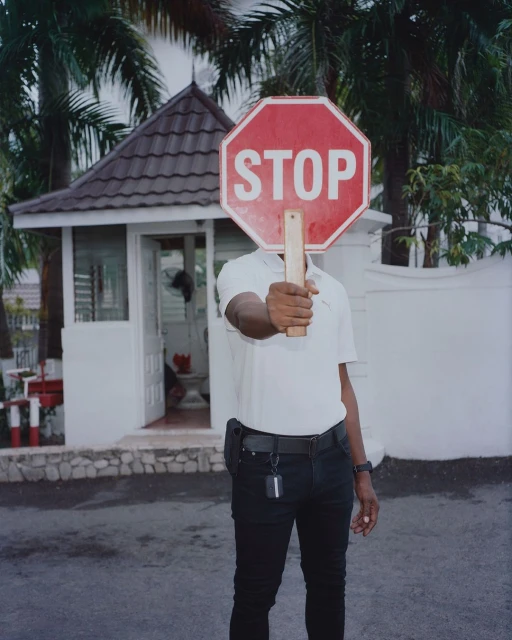 Person holds red stop sign with white letters in front of their face on an outdoor street near a white building and green trees.