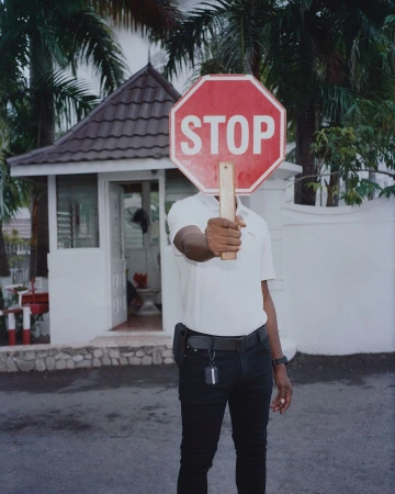 Person holds red stop sign with white letters in front of their face on an outdoor street near a white building and green trees.