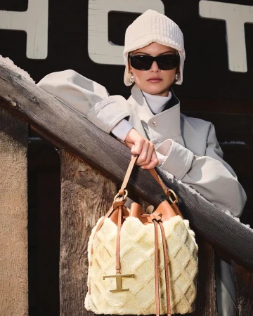 Model in cream winter hat and sunglasses leaning on a wooden railing holding a shearling bucket bag by Paolo Zerbini