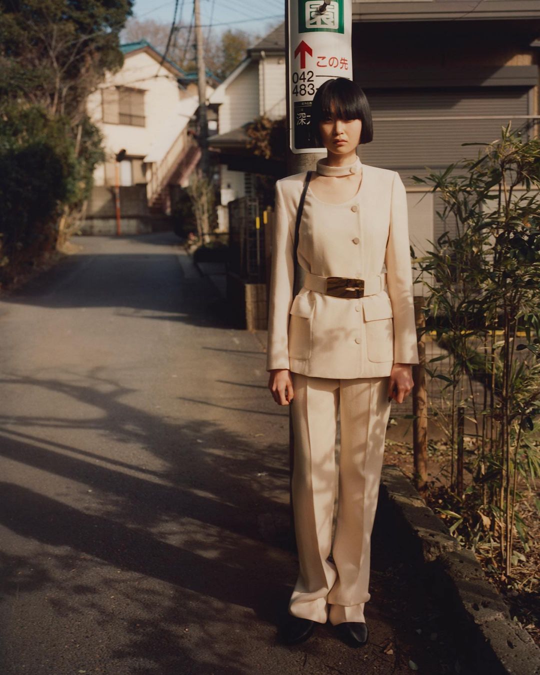 Woman in a beige suit stands on a street with a utility pole in the background, captured in natural light on a sunny day.