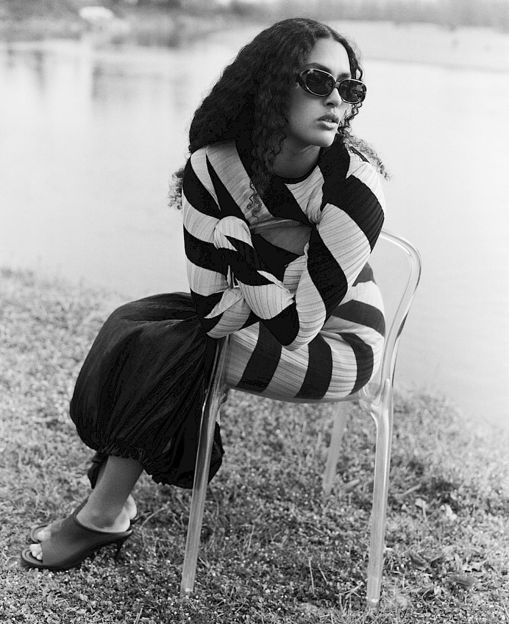 A young adult woman with curly hair sits on a transparent chair in a black and white fashion portrait, wearing sunglasses and a striped dress.