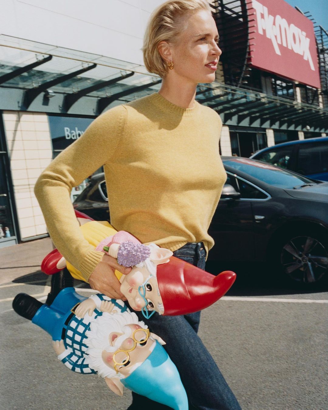 Woman in yellow sweater carries two garden gnomes in urban setting in front of a TK Maxx retail store on a sunny day.