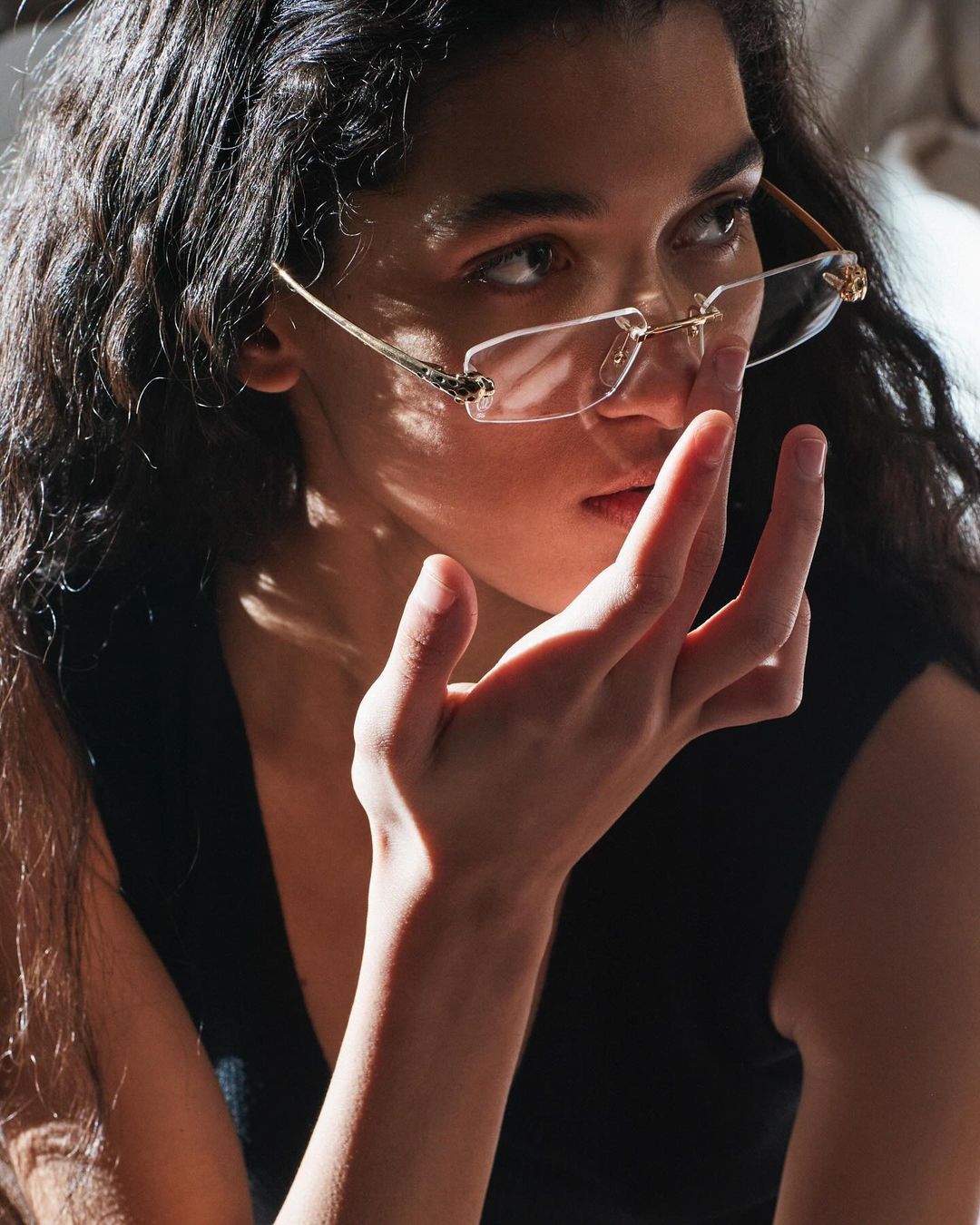 Close-up shot of an adult woman with wavy dark hair, wearing rimless glasses, face partially obscured by her hand.