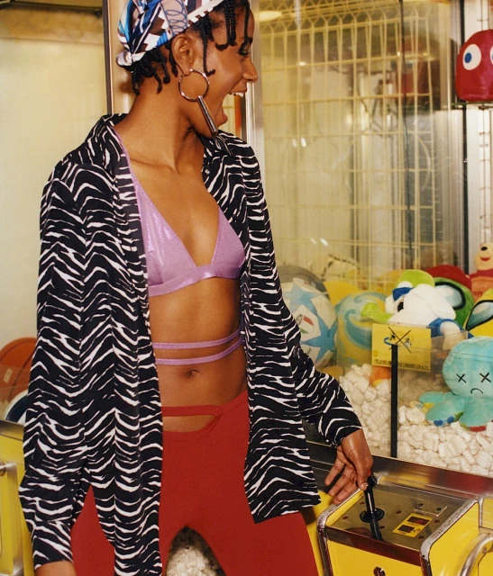 Stylish young woman poses near a claw machine wearing a zebra-print shirt, lavender crop top, red pants, and large hoop earrings.
